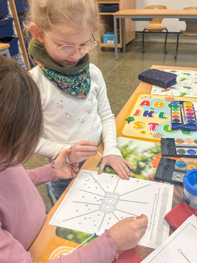 Zwei Kinder sitzen an einem Tisch und malen gemeinsam ein Ausmalbild mit einem Schmetterling und der Zahl 100.