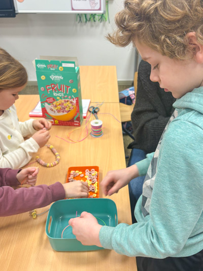 Kinder sitzen an einem Tisch und fädeln bunte Fruchtringe-Perlen auf Schnüre auf.