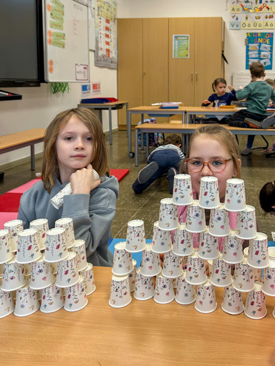 Zwei Kinder sitzen an einem Tisch mit mehreren Pyramiden aus Papierbechern im Klassenzimmer.