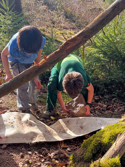 Zwei Jungen bücken sich im Wald und heben einen großen Stein neben einem umgestürzten Baumstamm.