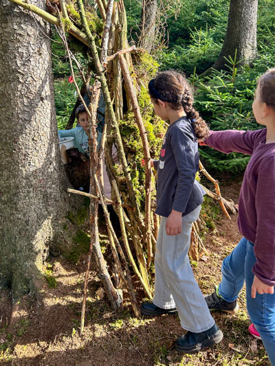 Drei Kinder stehen vor einer kleinen Hütte aus Ästen an einem Baum im Wald, ein Kind schaut aus der Hütte heraus.