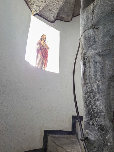 Statue einer heiligen Figur in einem hell erleuchteten Fenster über einer steinernen Wendeltreppe mit Metallgeländer.