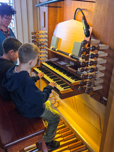 Zwei Jungen sitzen an einer Orgel mit mehreren Manualen, Registerzügen und Pedalen, eine Frau steht daneben.