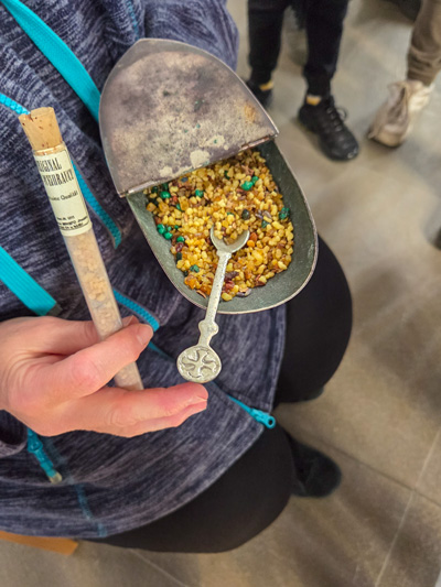 Hand hält eine kleine Metallpfanne mit buntem Granulat und einem kleinen Löffel mit verziertem Griff.