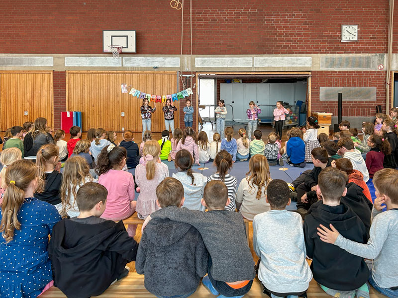 Kinder sitzen auf dem Boden einer Turnhalle und schauen einer Gruppe von Kindern zu, die vor einer Holzwand mit Basketballkorb und bunter Girlande auftreten.