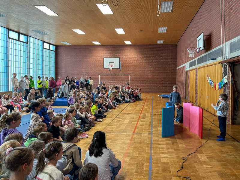 Kinder sitzen auf dem Boden und auf Matten in einer Turnhalle und schauen drei Kindern zu, die an einem Mikrofon vor bunten Stellwänden sprechen.