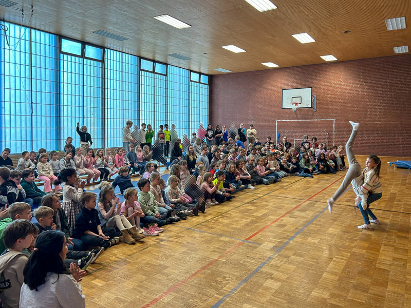 Kinder sitzen auf dem Boden einer Turnhalle und schauen zwei Mädchen zu, von denen eines einen Handstand macht.
