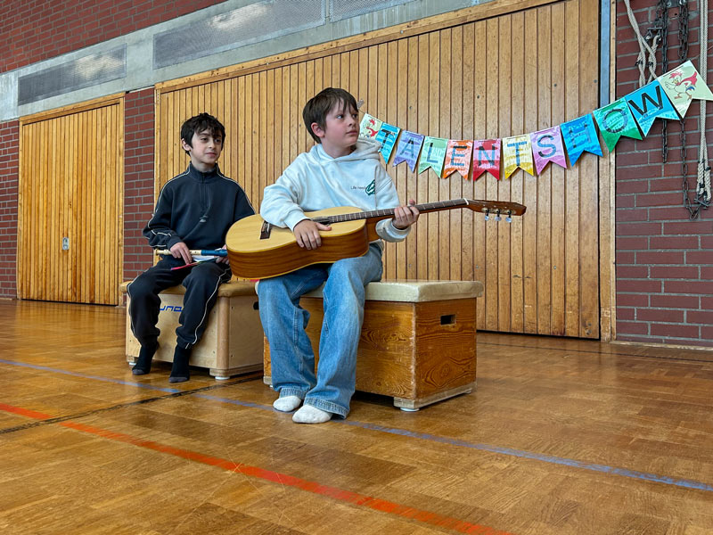 Zwei Kinder sitzen auf Holzkisten in einer Turnhalle, eines spielt Gitarre, im Hintergrund hängt ein bunter Schriftzug „TALENTSHOW“.
