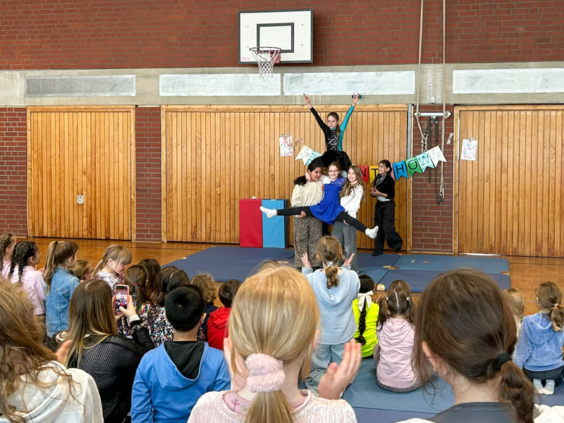 Kinder führen in einer Turnhalle eine Akrobatik-Übung vor einem Publikum auf, ein Mädchen steht auf den Schultern von zwei anderen Kindern und streckt die Arme nach oben.