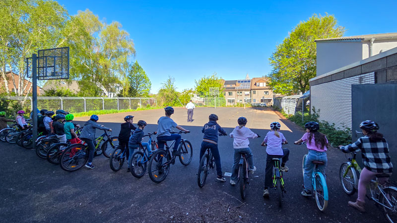 Gruppe von Kindern mit Fahrradhelmen auf Fahrrädern steht auf einem Schulhof und blickt zu einer Person in der Ferne.