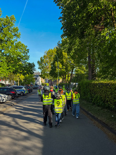 Gruppe von Kindern mit gelben Warnwesten und Helmen geht auf einer von Bäumen gesäumten Straße.