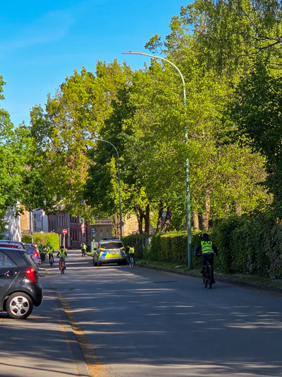 Straße mit parkenden Autos, mehreren Radfahrern und einem Polizeiauto, umgeben von Bäumen und Häusern.
