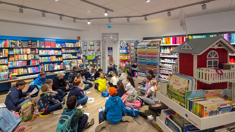 Kinder sitzen im Kreis auf dem Boden eines Buchladens und hören einer vorlesenden Frau zu.