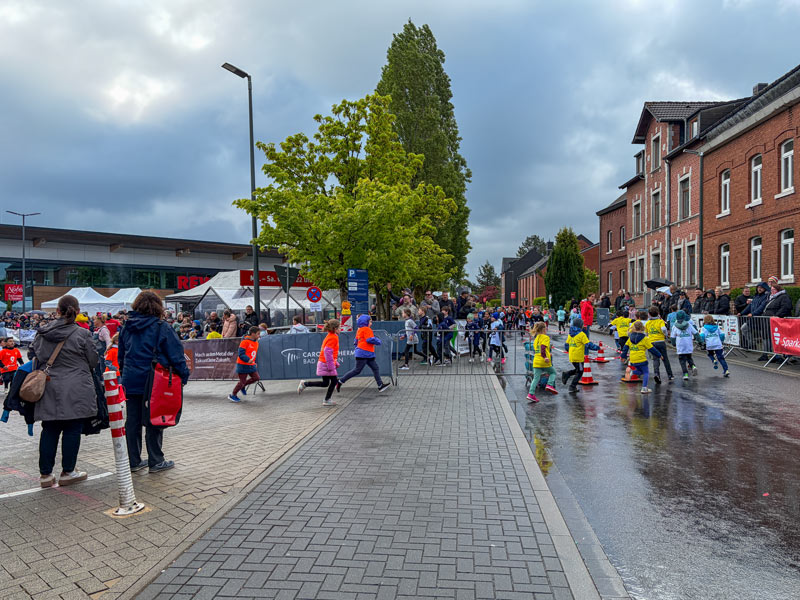 Kinder in bunten T-Shirts laufen auf einer nassen Straße, umgeben von Zuschauern und Gebäuden bei bewölktem Himmel.