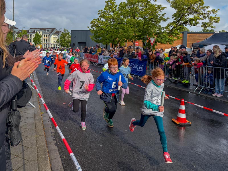 Kinder in Sportkleidung laufen auf einer abgesperrten Straße bei einem Laufwettbewerb, Zuschauer klatschen am Straßenrand.