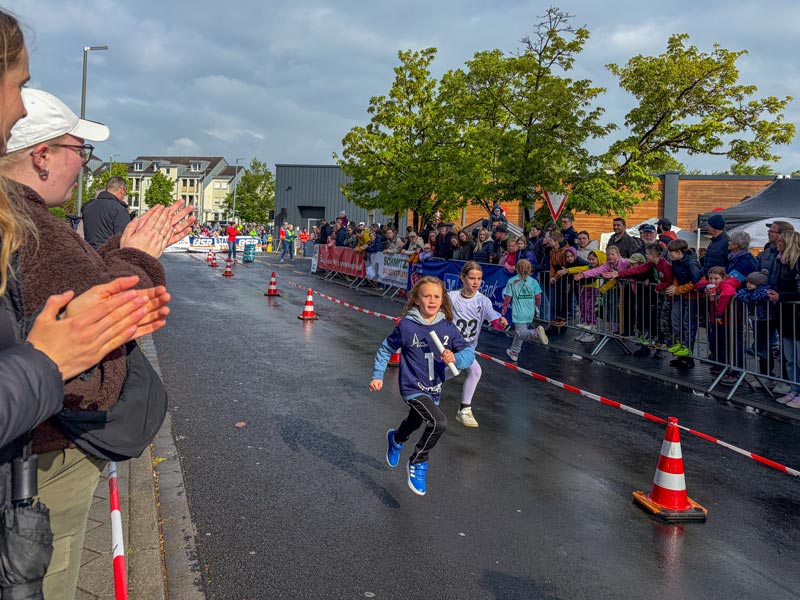 Zwei Kinder laufen auf einer abgesperrten Straße, links applaudieren Zuschauer, rechts stehen weitere Zuschauer hinter Absperrungen.