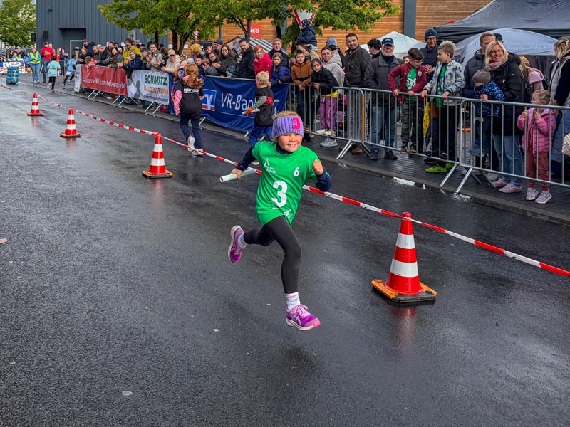 Kind in grüner Sportkleidung läuft auf nasser Straße an Absperrung mit Zuschauern vorbei.