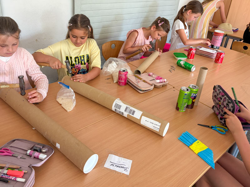 Kinder sitzen an einem Tisch und bearbeiten lange braune Papprollen mit Werkzeugen und Bastelmaterialien.