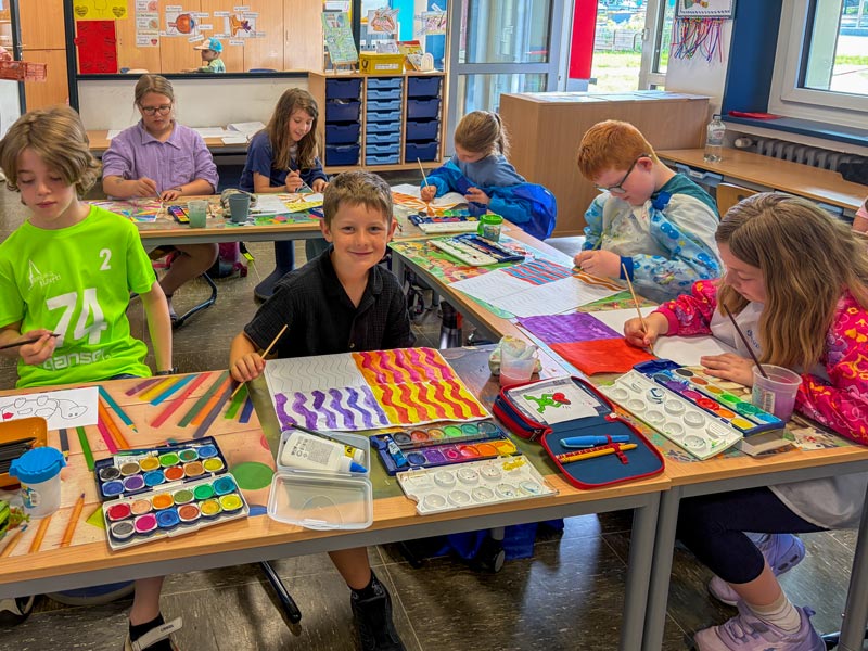 Kinder sitzen an Tischen in einem Klassenraum und malen mit Wasserfarben auf Papier.