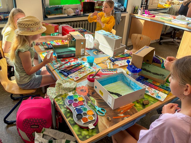 Kinder sitzen an einem Tisch mit Bastelmaterialien und bemalten Kartons in einem Klassenraum.