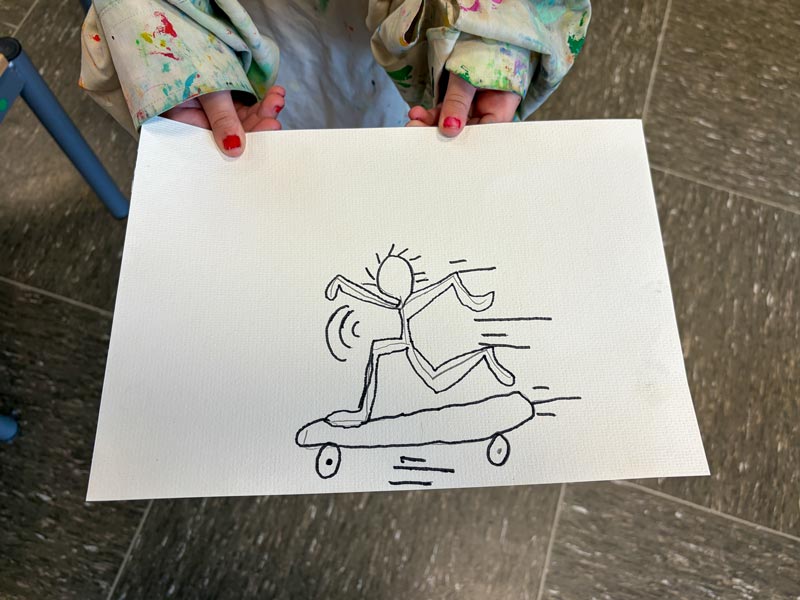 Kind hält ein Blatt mit einer Strichmännchenzeichnung, das auf einem Skateboard fährt.