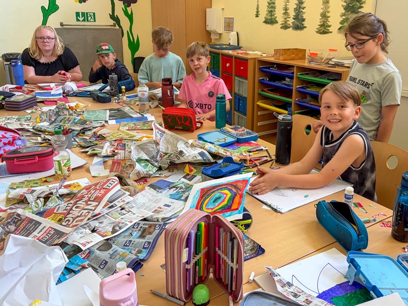 Kinder sitzen an einem Tisch mit vielen Bastelmaterialien, Zeitungen und Stiften in einem Klassenraum.