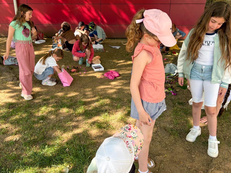Mehrere Kinder sitzen auf einer Wiese vor einer roten Wand, einige bücken sich und andere stehen.