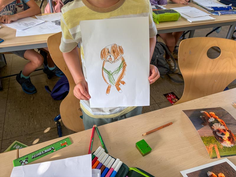 Kind hält selbstgemalte Zeichnung eines braun-weißen Hundes mit grünem Halsband in einem Klassenzimmer.