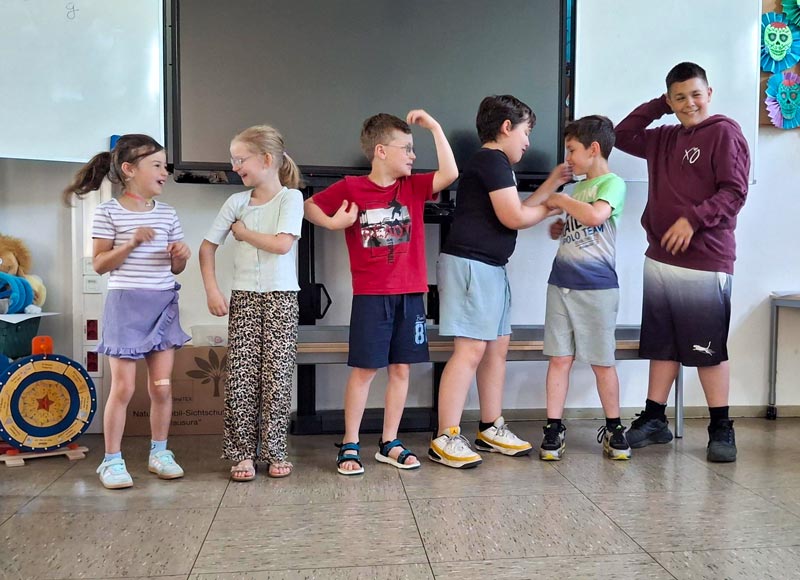 Sechs Kinder stehen nebeneinander in einem Klassenraum, einige mit erhobenen Armen und unterschiedliche Kleidung tragend.