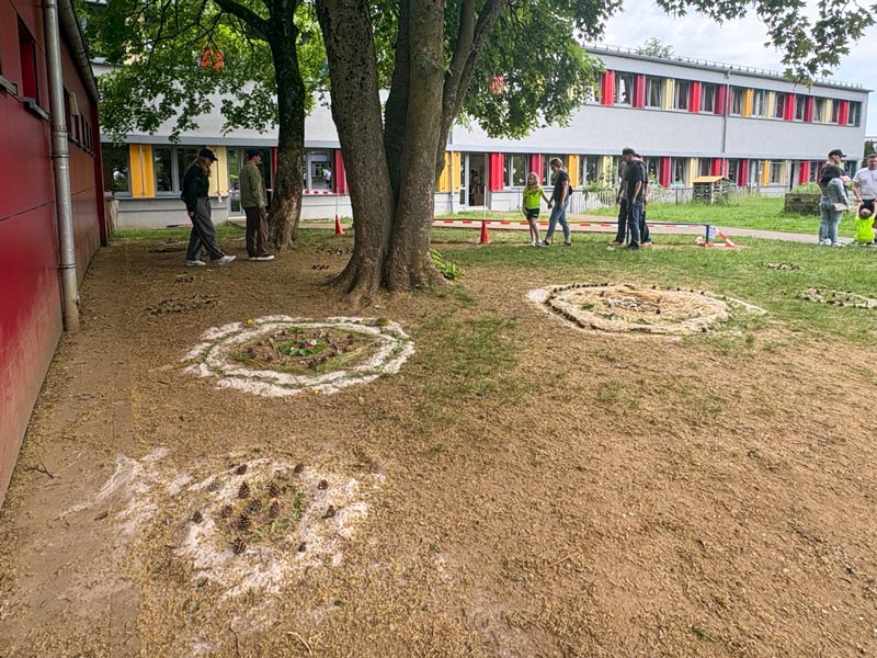 Schulhof mit mehreren kreisförmigen Beeten auf dem Boden, umgeben von Bäumen und mehreren Personen vor einem mehrstöckigen Schulgebäude mit bunten Fensterläden.