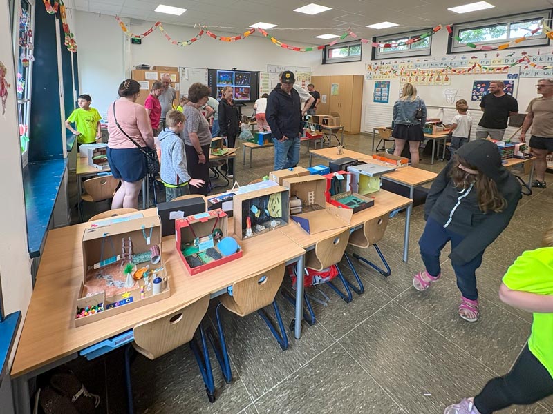 Klassenraum mit mehreren Tischen, auf denen Bastelarbeiten aus Pappkartons stehen, und Menschen, die sich die Arbeiten ansehen.