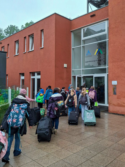 Gruppe von Kindern mit Rucksäcken und Rollkoffern betritt ein rotes Gebäude mit Glasfassade und Logo an der Eingangstür.