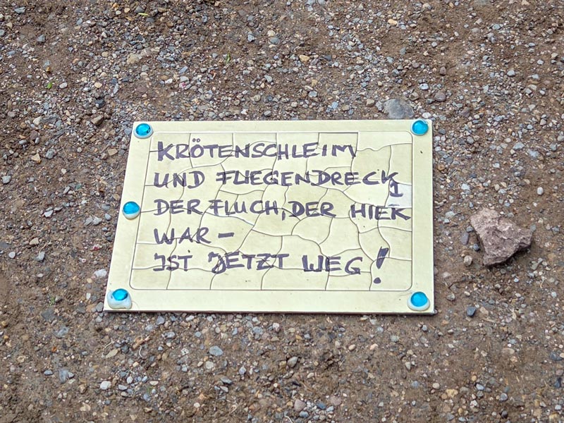 Handgeschriebene Tafel mit Text auf kiesigem Boden befestigt durch vier blaue Nägel.