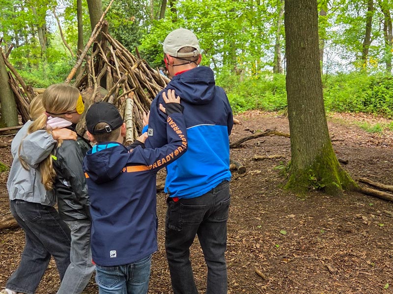 Vier Kinder mit verbundenen Augen tasten sich im Wald an einem Baumstamm entlang.