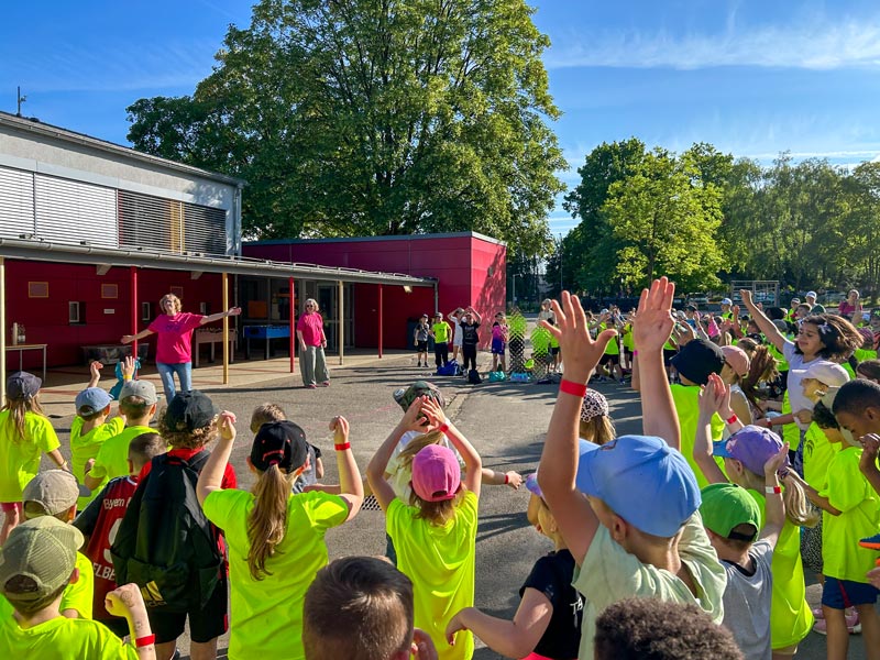 Kinder in neongelben T-Shirts und bunten Mützen stehen im Freien vor einem roten Gebäude und heben die Arme, während zwei Erwachsene ihnen gegenüberstehen.