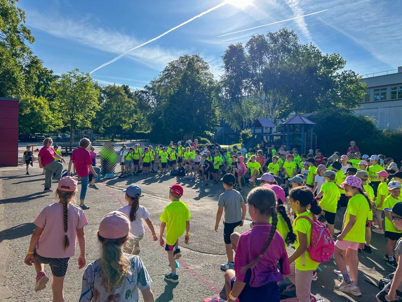 Viele Kinder in neongelben T-Shirts und einige Erwachsene versammeln sich auf einem Schulhof bei sonnigem Wetter mit Bäumen und Spielgeräten im Hintergrund.