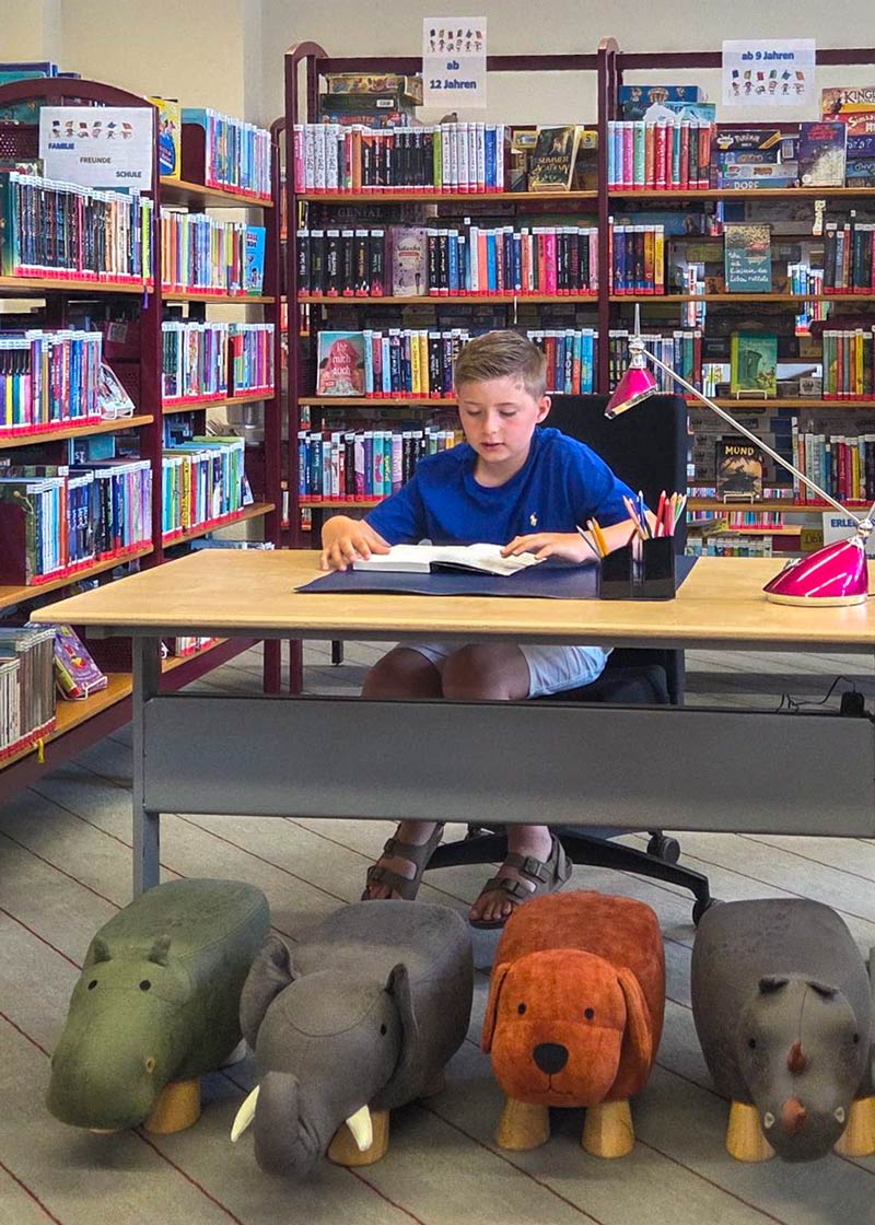 Kind sitzt an einem Tisch in einer Bibliothek vor einem aufgeschlagenen Buch, unter dem Tisch stehen vier Tierhocker in Form eines Nilpferds, Elefanten, Hundes und Nashorns.