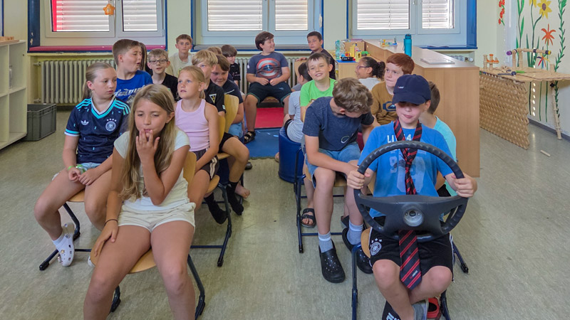 Gruppe von Kindern sitzt in einem Klassenzimmer, ein Kind hält ein Lenkrad und trägt Krawatte.
