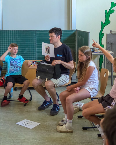Mehrere Kinder sitzen im Stuhlkreis in einem Klassenzimmer, eine Person hält eine Karte mit einem durchgestrichenen Symbol hoch.