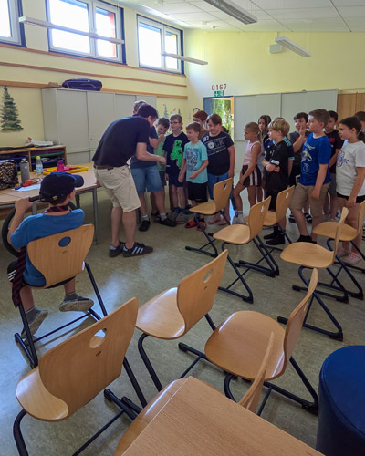 Gruppe von Kindern steht in einem Klassenraum vor einem Erwachsenen, mehrere leere Stühle im Vordergrund.