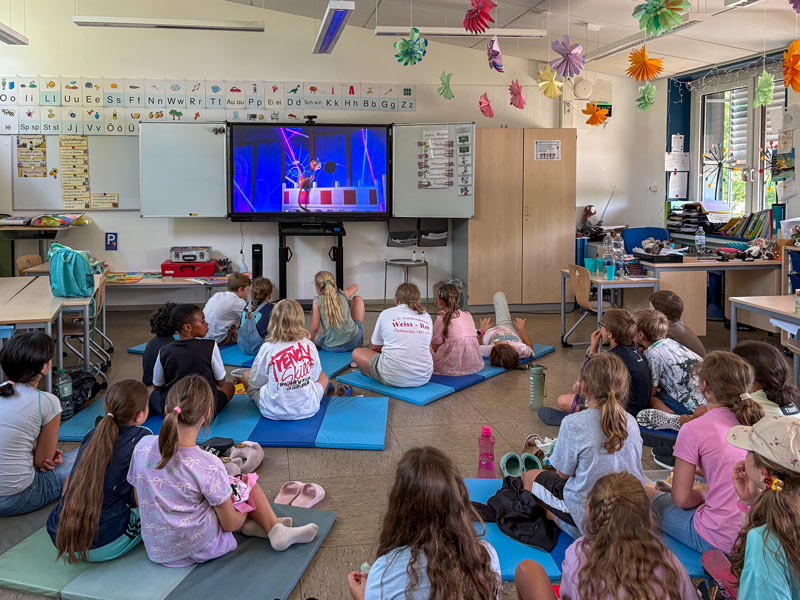 Kinder sitzen auf Matten in einem Klassenzimmer und schauen auf einen Bildschirm an der Wand.