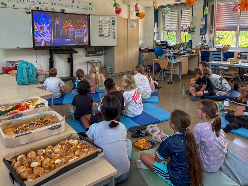 Kinder sitzen auf blauen Matten in einem Klassenzimmer und schauen auf einen Bildschirm an der Wand. Auf einem Tisch stehen Behälter mit Snacks.
