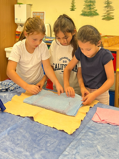 Drei Mädchen falten gemeinsam ein blau-weiß gestreiftes Tuch auf einem Tisch mit blauer Decke.