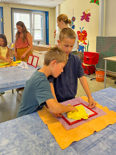 Zwei Jungen falten gelbe Stoffstücke auf einem Tisch mit orangem und rot-weiß kariertem Tuch im Klassenraum mit bunten Schmetterlingsdekorationen an der Wand.