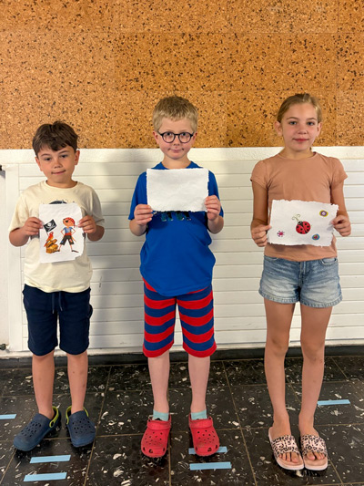 Drei Kinder stehen nebeneinander vor einer Wand und halten jeweils ein selbstgestaltetes Stück Papier hoch.
