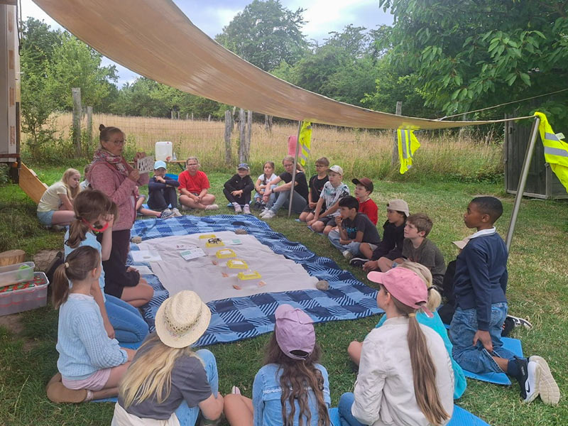 Kinder sitzen im Kreis auf einer Wiese unter einem Sonnensegel und hören einer stehenden Frau zu.