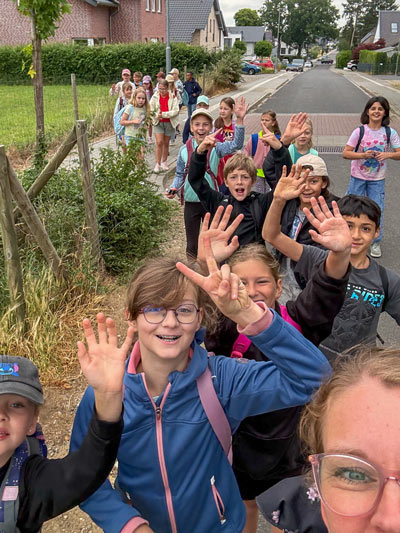 Gruppe von Kindern mit Rucksäcken, die auf einem Gehweg stehen und in die Kamera winken.