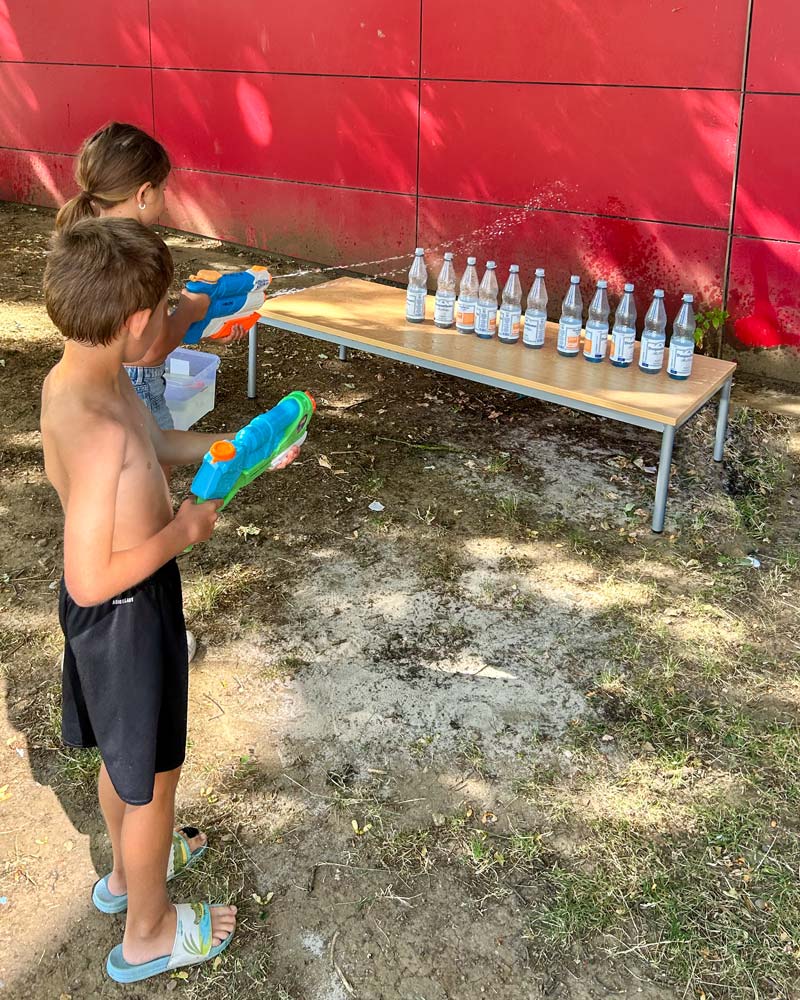 Zwei Kinder mit Wasserpistolen zielen auf eine Reihe von Plastikflaschen auf einem Tisch vor einer roten Wand.