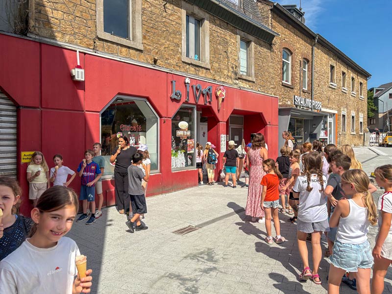 Kinder stehen in einer Schlange vor einem roten Gebäude mit der Aufschrift „Liva“ und essen Eis.
