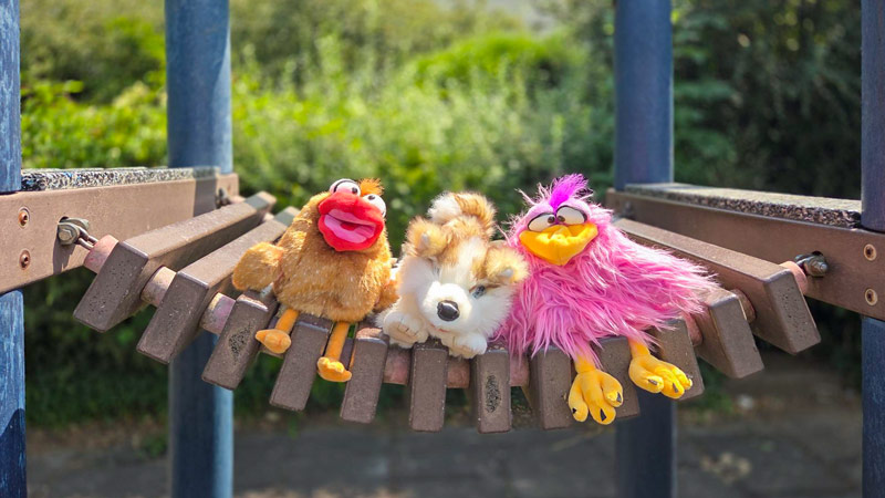 Drei bunte Plüschtiere sitzen nebeneinander auf einer hölzernen Hängematte im Freien.
